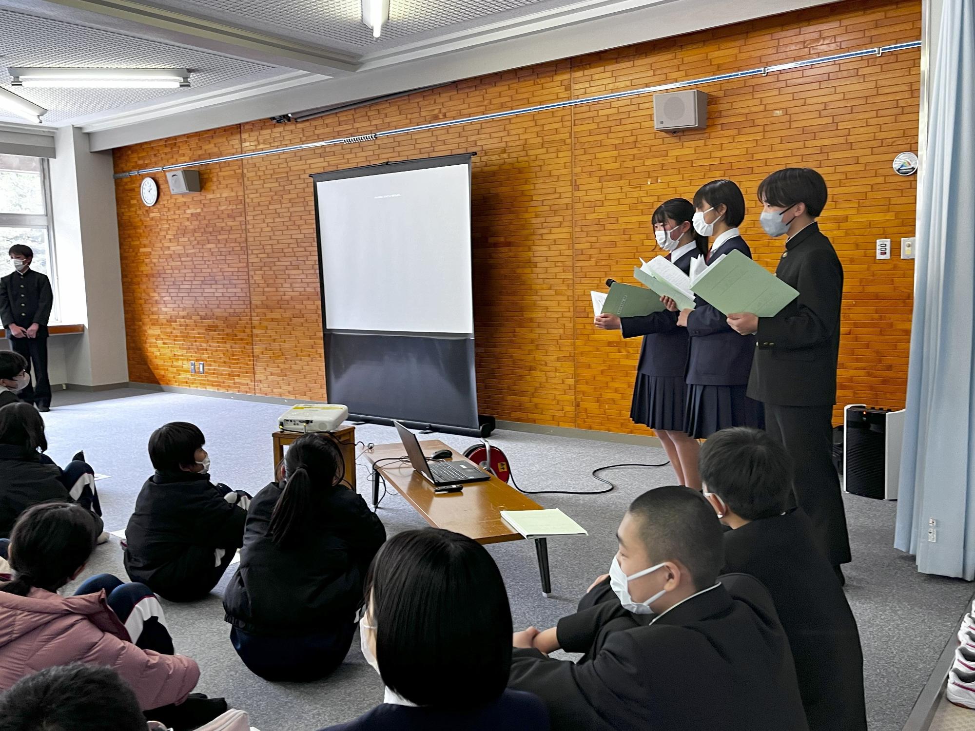 生徒たちが前に立ち、資料を手に発表しており、それを座って聞く生徒たちの写真