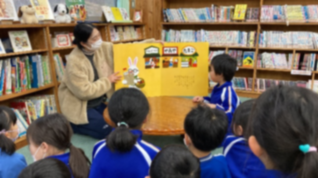 図書室で教師が紙芝居を使って児童たちに読み聞かせをしている写真