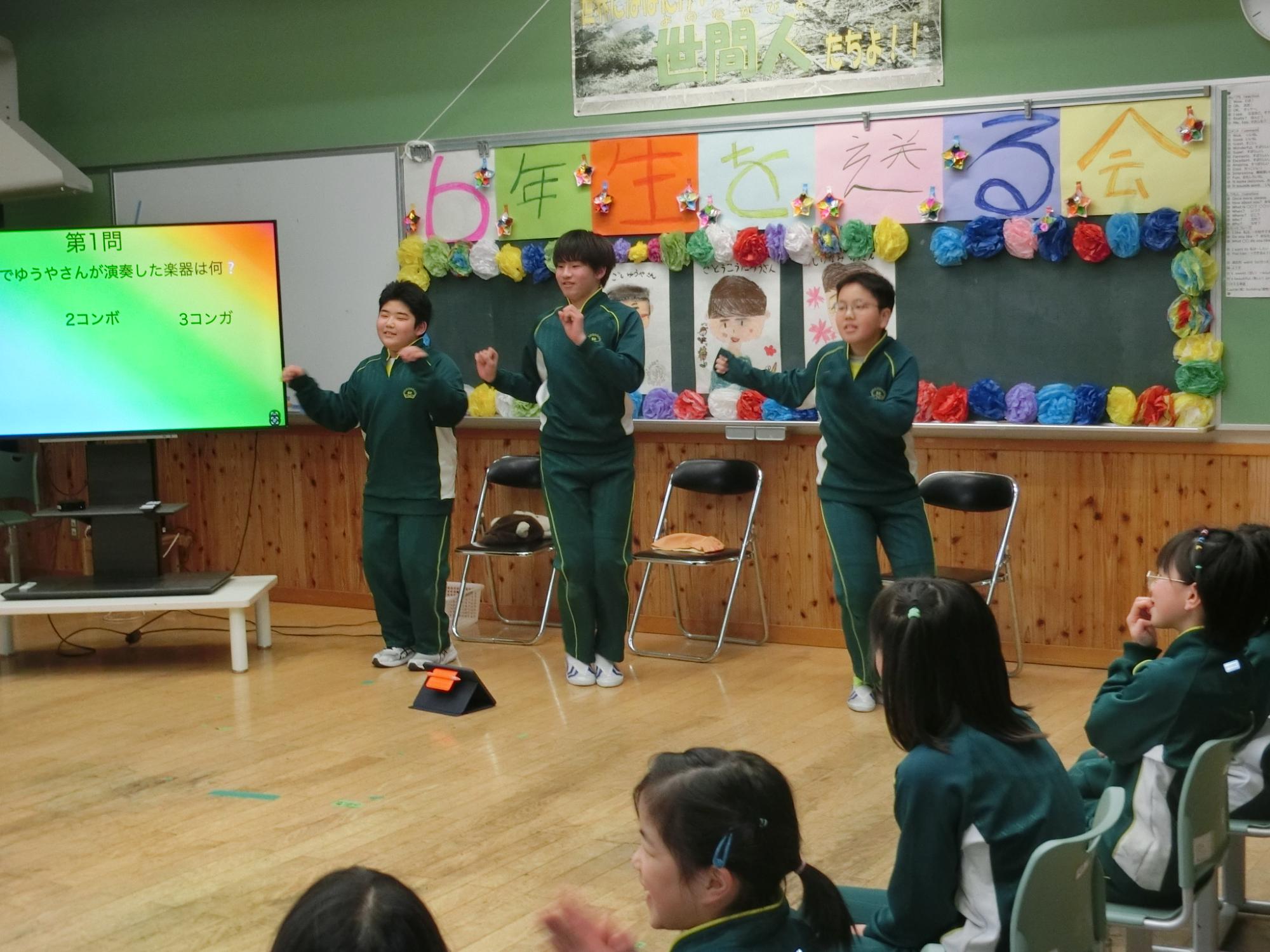 飾り付けられた黒板の前で出し物を披露する3人の生徒と席につき手を叩いたりしながら見守る生徒達が写った6年生を送る会の写真
