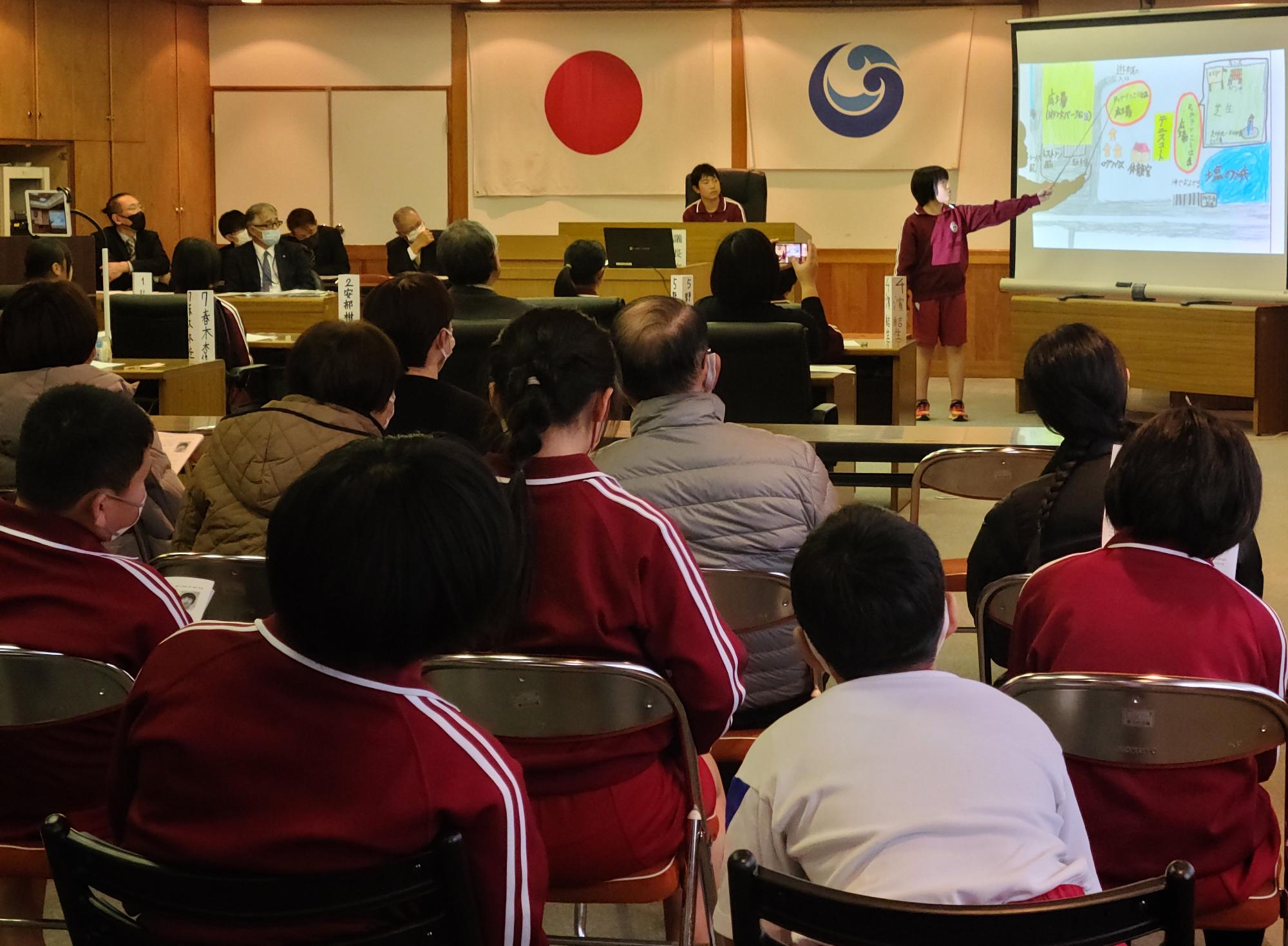 公民館で児童が前に立って発表を行い、他の児童や大人が座りながら見聞きしている写真