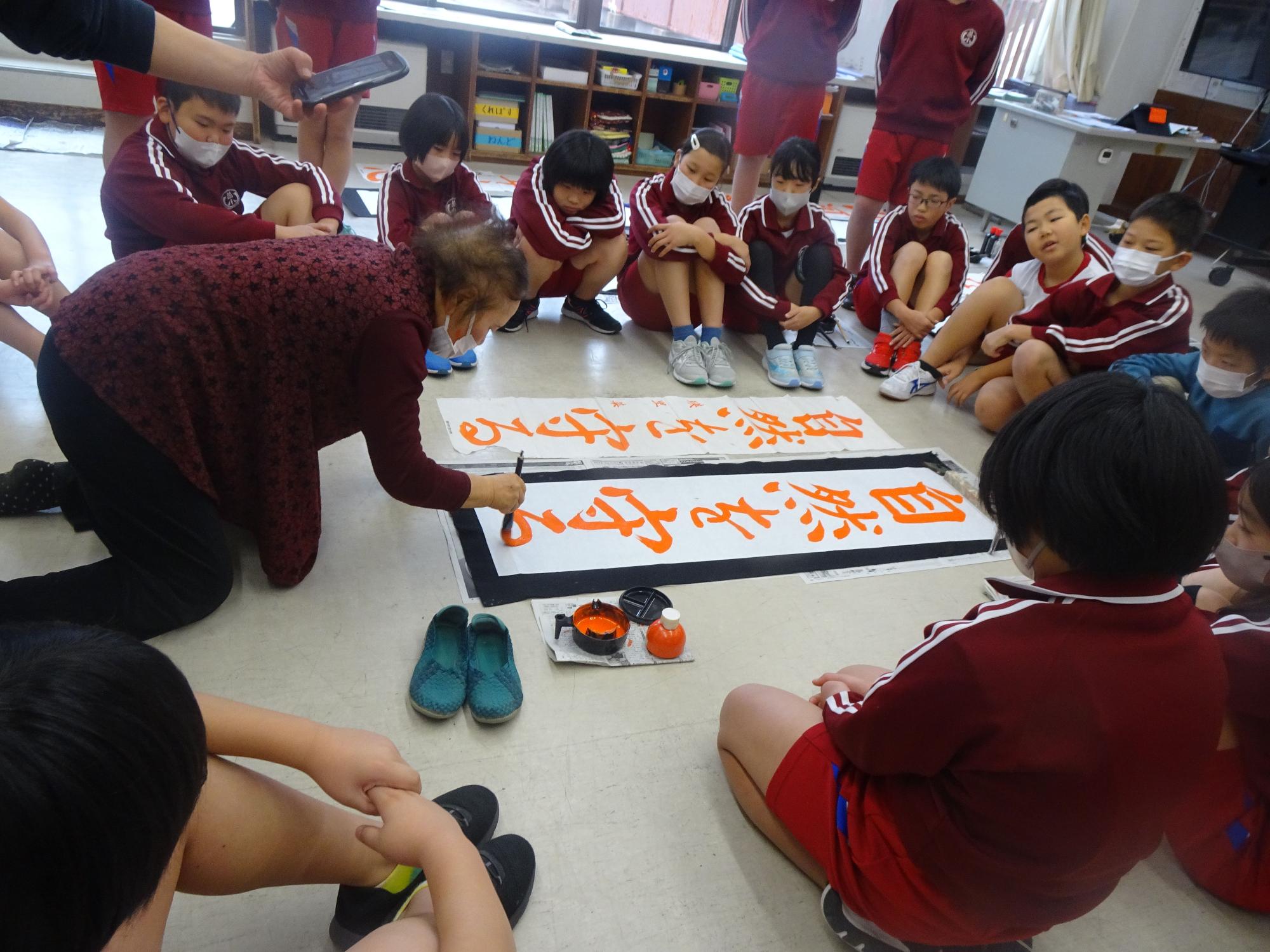 教室で児童たちが輪になって座り、講師が書道を教えている写真