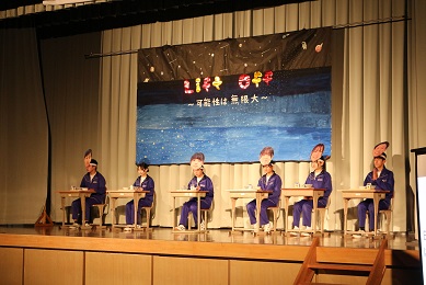 舞台上で椅子に座った作業着姿の生徒たちが発表をしている写真