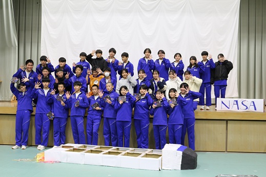 体育館の舞台上で青いジャージを着た生徒たちが集合写真を撮っている写真