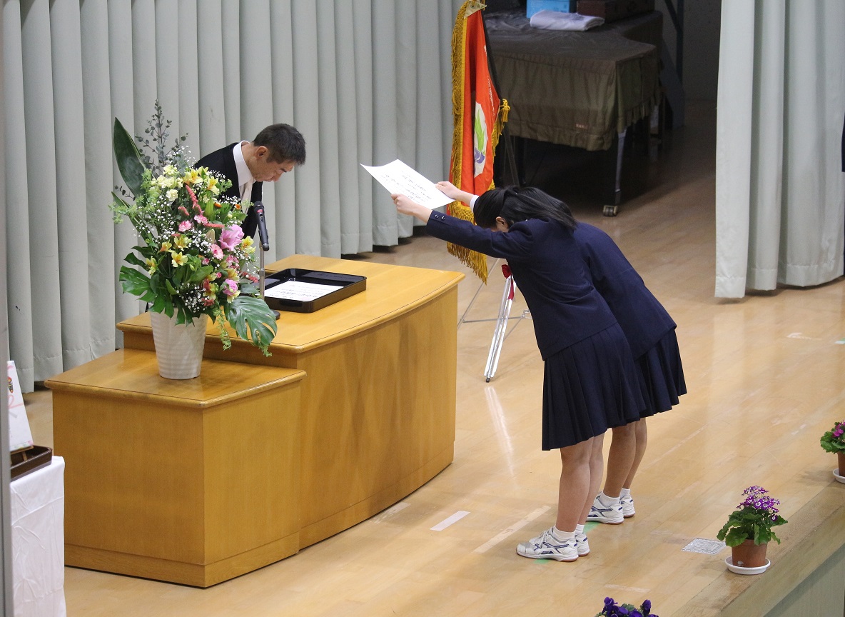 式典の壇上で女子生徒が卒業証書を受け取っている写真