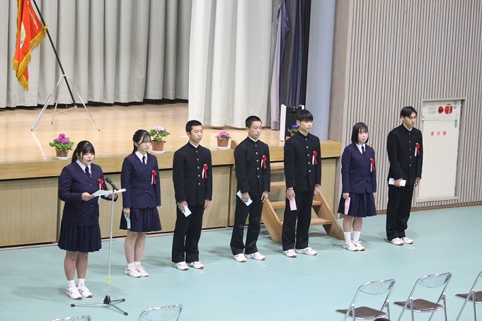 体育館の舞台前で生徒代表が整列してあいさつをしている写真