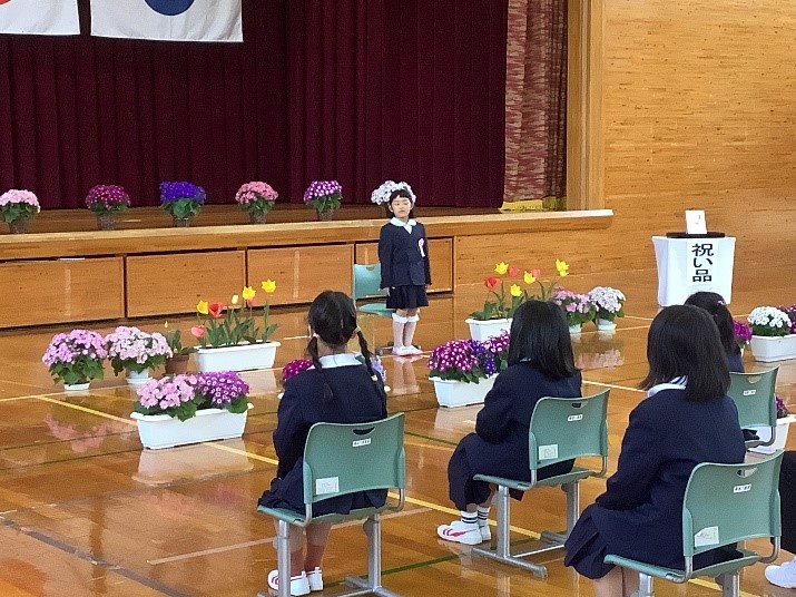 お祝いのお花に囲まれ、1人でも堂々と前に立ち在校生の方を見ている新1年生の写真