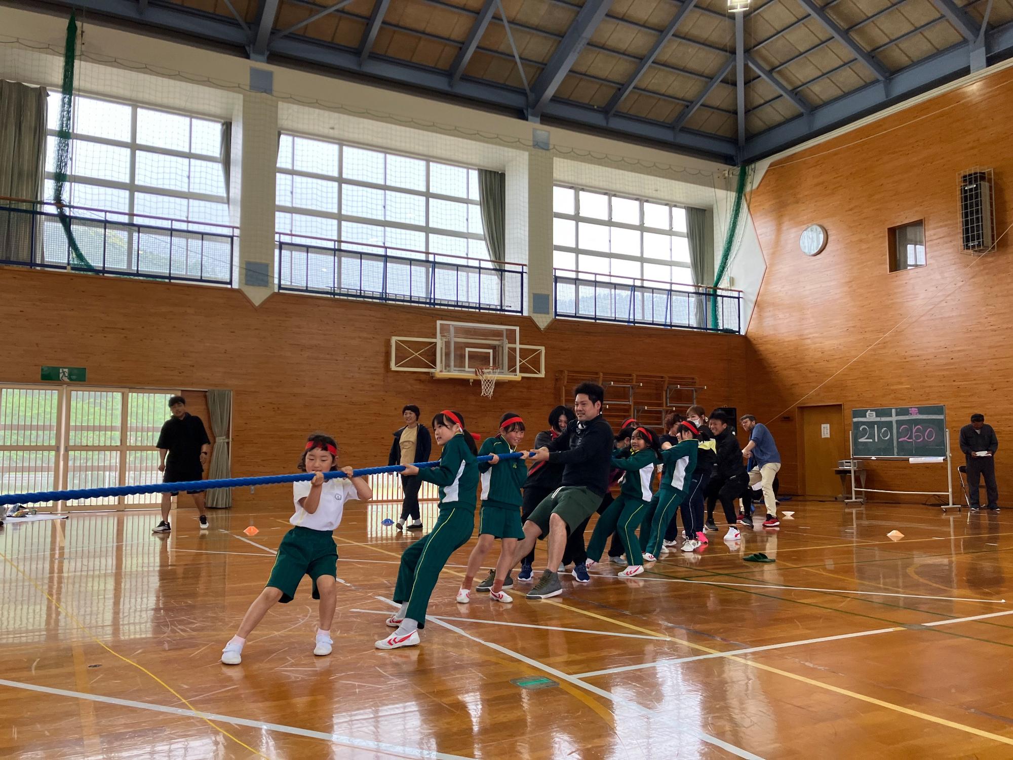 頭に赤い鉢巻をまいた生徒達と一緒になって綱を引いている数人の大人達と 赤組白組の点数を書いた得点板の写真