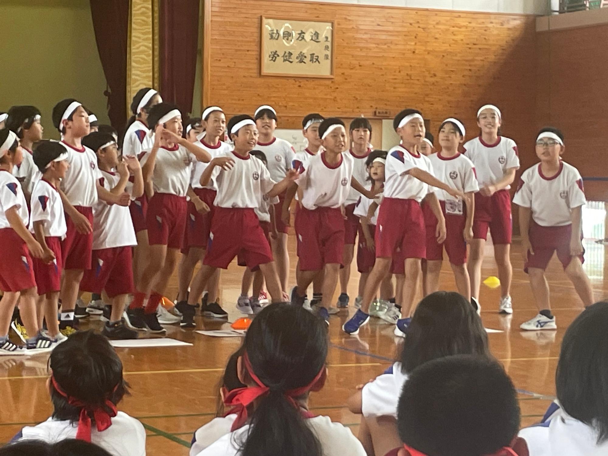 体育館で体育着と白色のハチマキを着用した数十人の児童たちが団体演技をしている写真