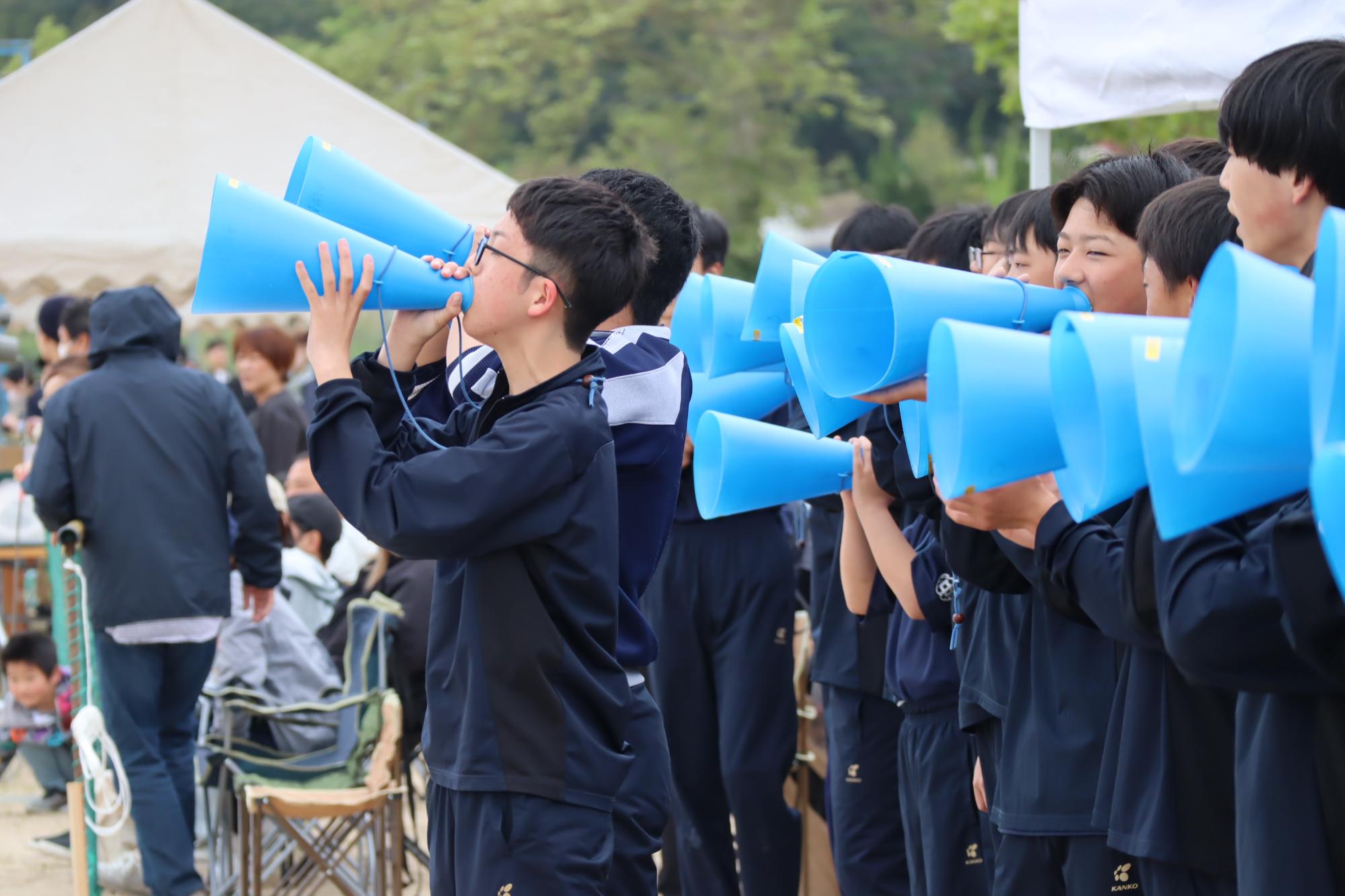 競技場で生徒たちが青いメガホンで声援を送っている写真