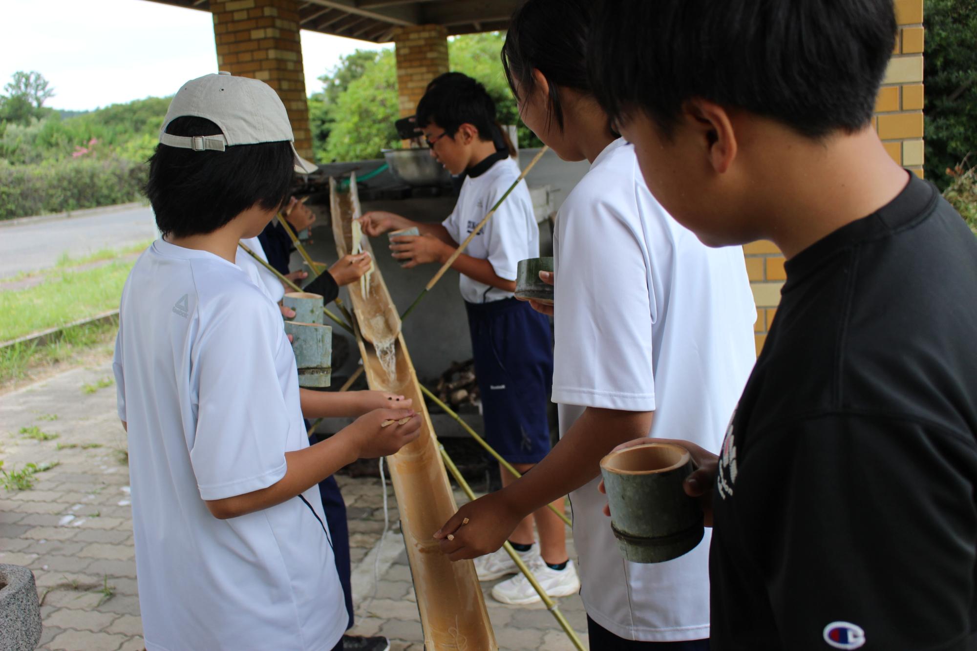 屋外の屋根付きスペースで竹を使った流しそうめんを楽しむ生徒たちの写真