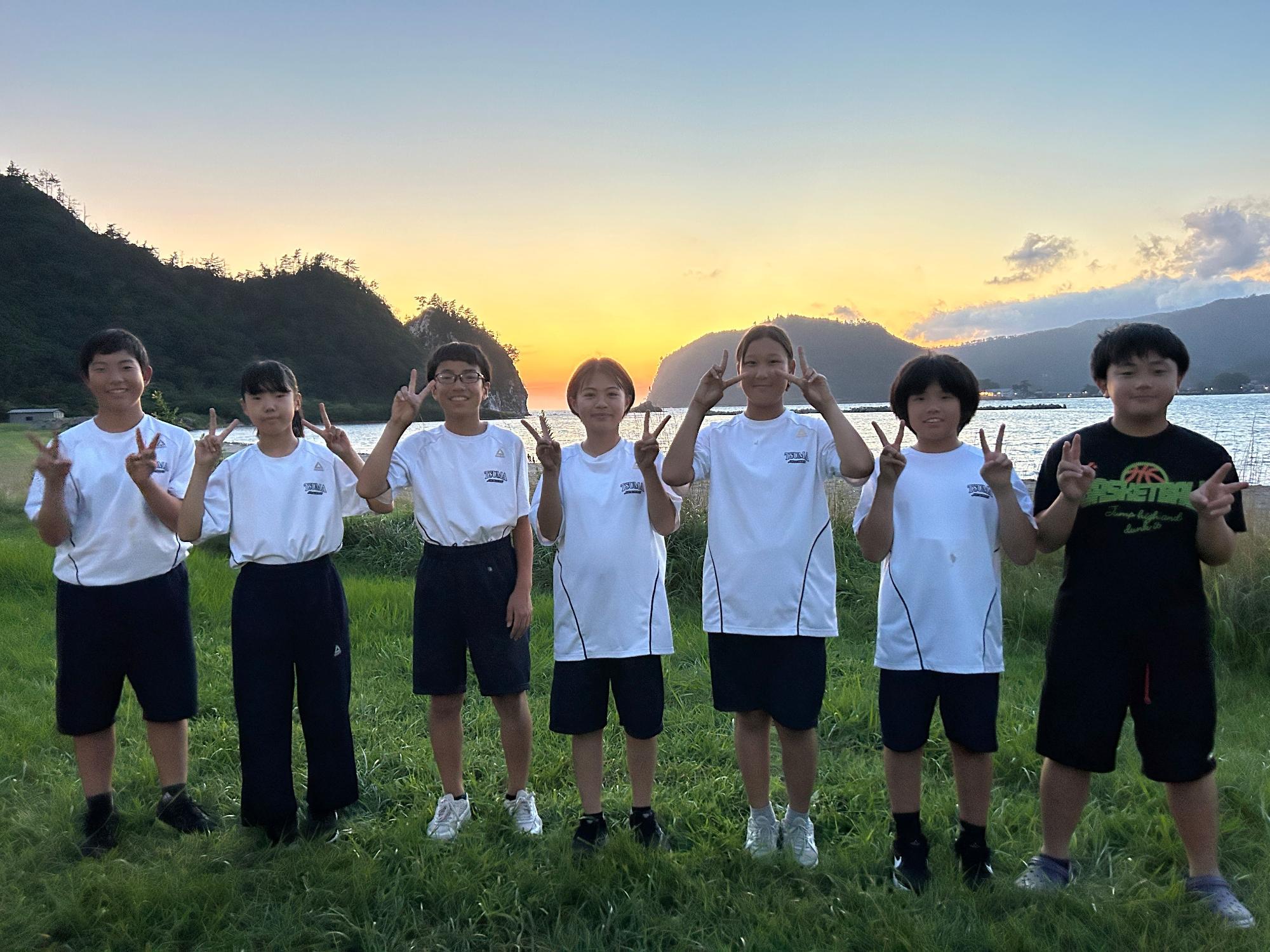 海辺の草地で夕焼けを背景に並んでポーズをとる生徒たちの写真