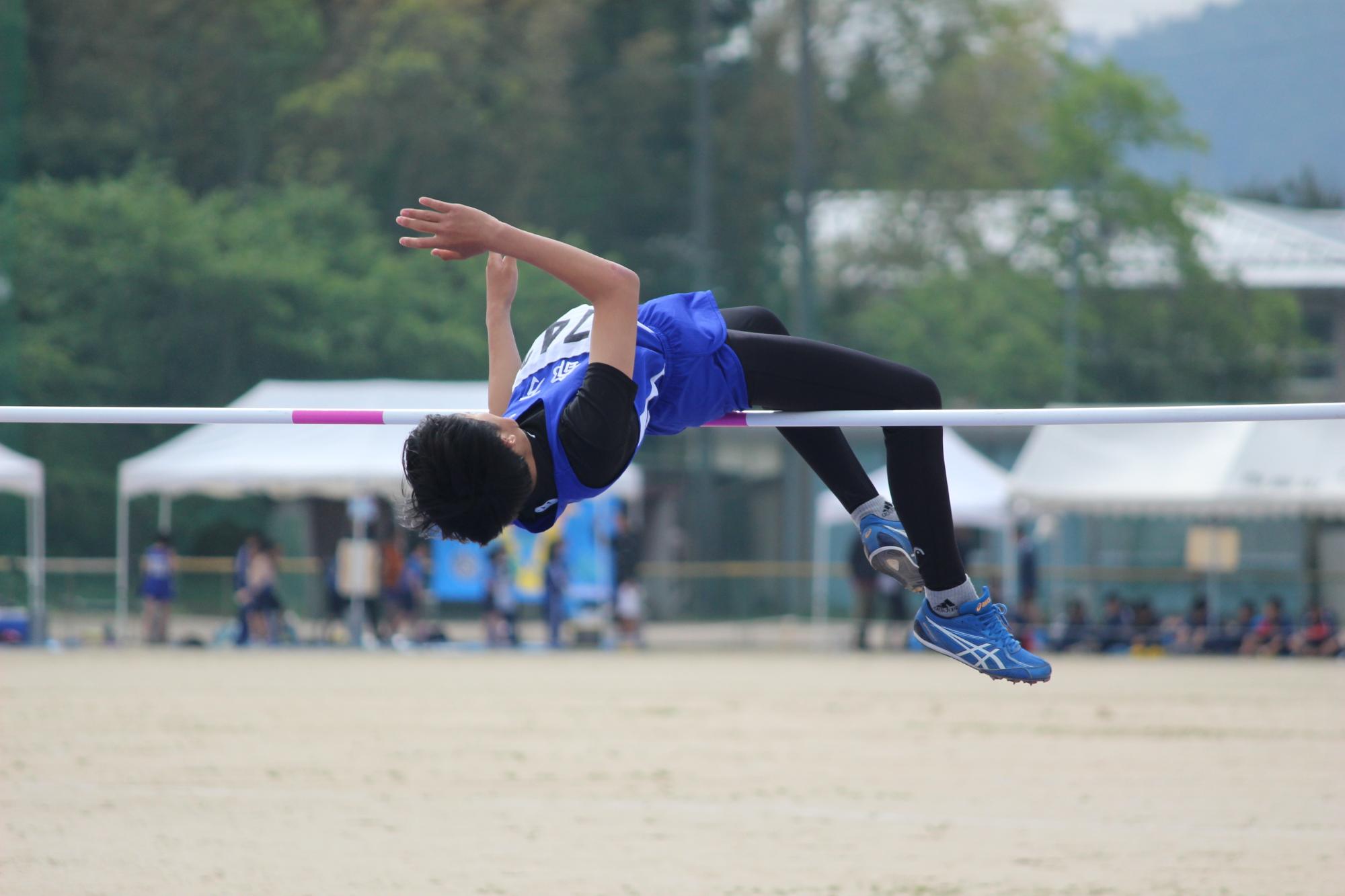 校庭で一人の生徒が背面跳びで白色のバーを跳び越える瞬間の写真