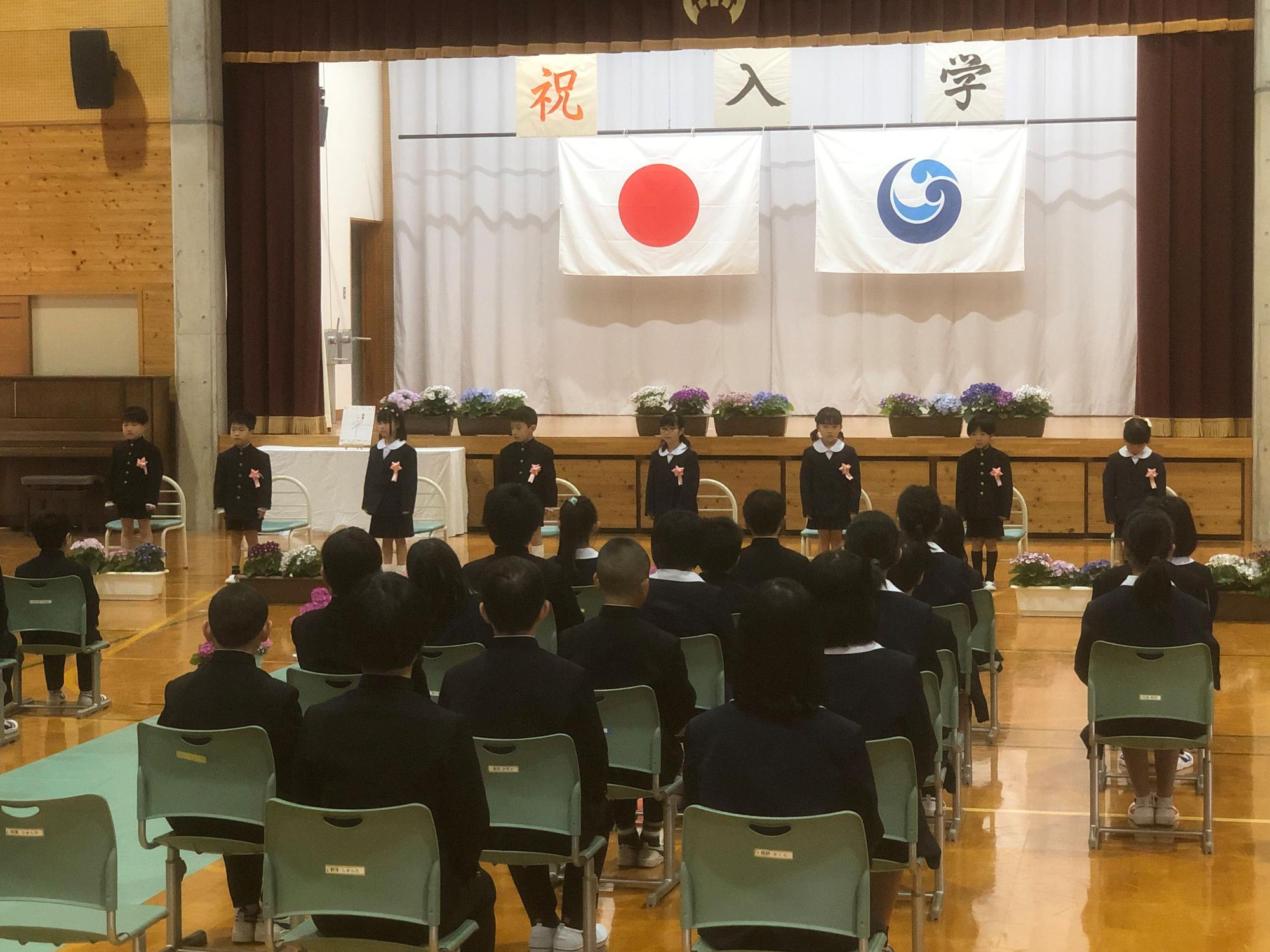 体育館で新入生が舞台前に整列している児童と椅子に座っている児童の後ろ姿の写真