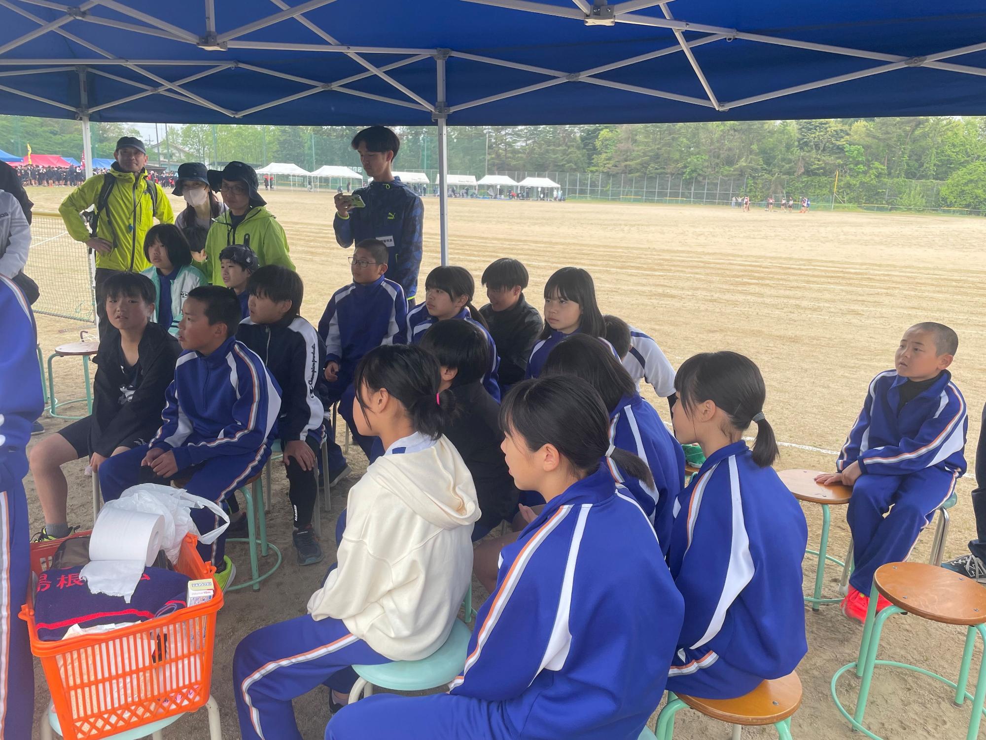 運動場のテントの下で児童が話を聞いている写真