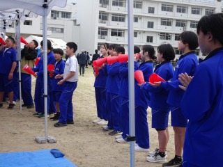 校庭のテント下で青いジャージを着た生徒たちが声援を送っている写真