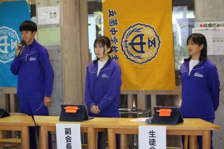 学校のホールで三人の生徒が前に立ってあいさつをしている写真