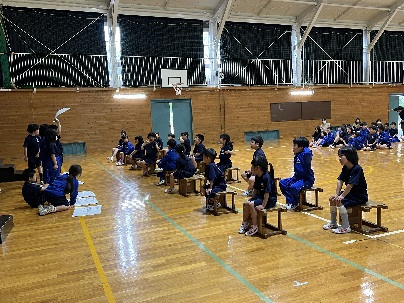 体育館で青い体操服を着た児童たちが舞台前で発表を見ている写真