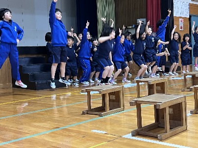 体育館の舞台で青い体操服を着た児童たちが元気にジャンプして発表している写真