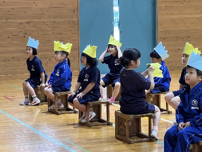 体育館で黄色と水色の王冠をかぶった児童たちが椅子に座って発表を見ている写真