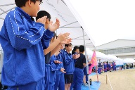 青いジャージを着た児童たちがテントの下で拍手をしながら応援している写真