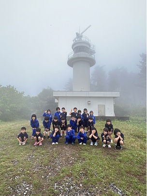 霧の立ちこめる山頂で青い体操服を着た児童たちが灯台の前に並んで記念撮影をしている写真