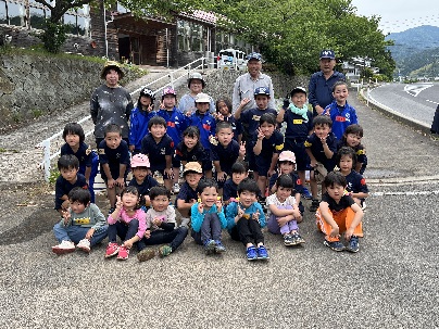 校舎前の坂道で帽子をかぶった児童たちと地域の方々が並んで集合写真を撮っている写真