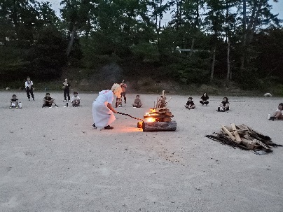 夕方のキャンプ場で子どもたちが見守る中、リーダーがキャンプファイヤーに火をつけている写真