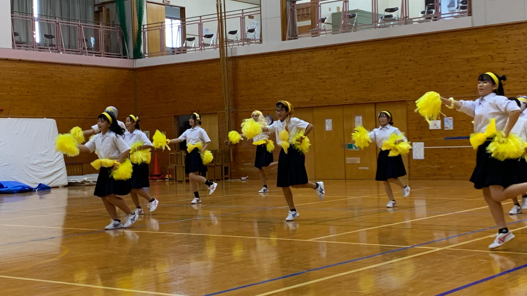 体育館で黄色いポンポンを持った生徒たちがダンスを披露している写真