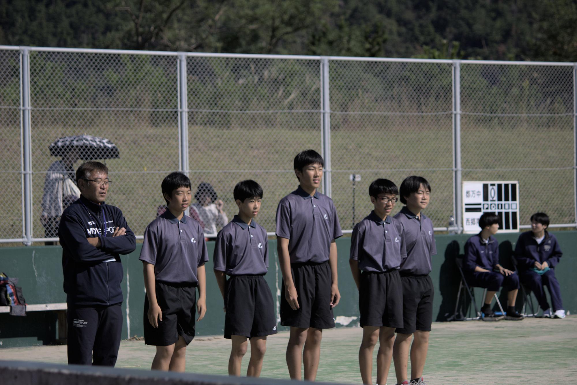 テニスコートで顧問の先生と男子生徒たちが整列している写真
