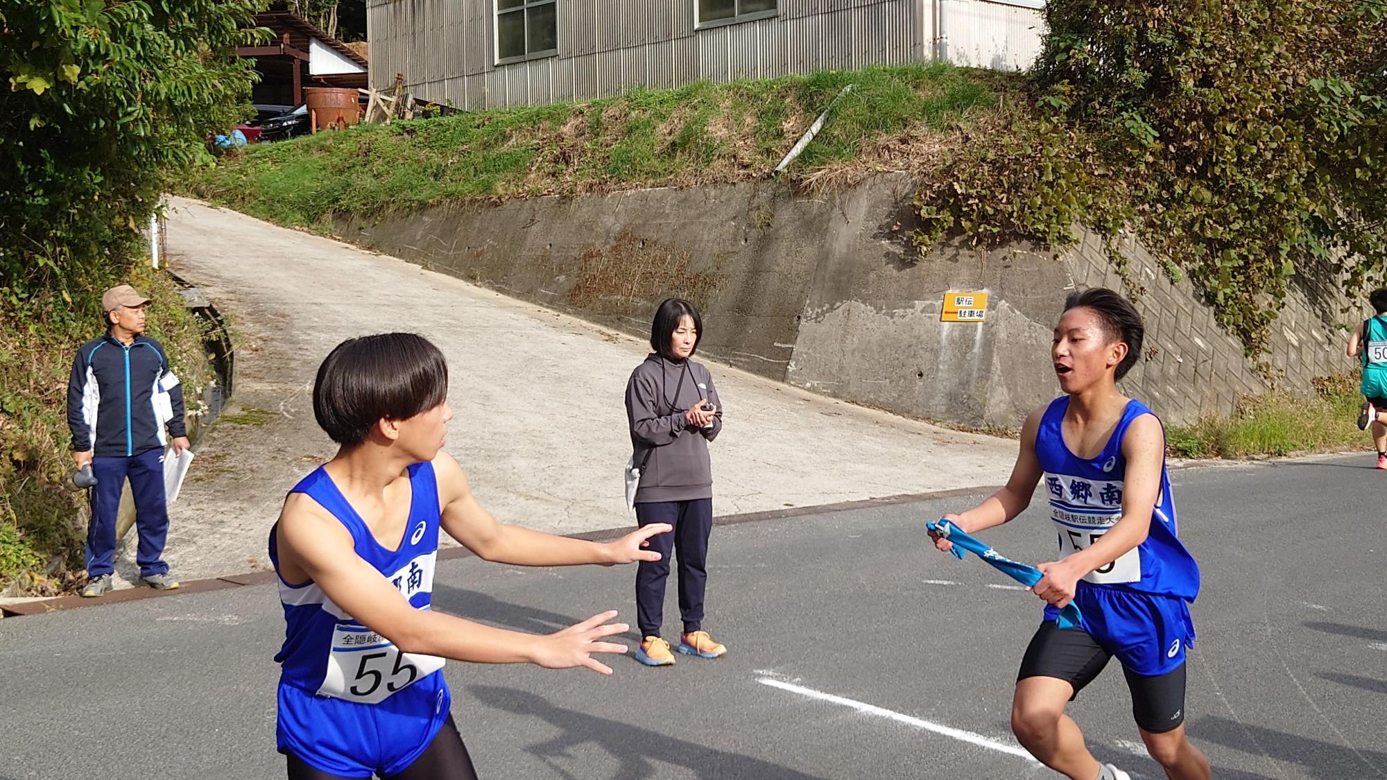 道路で男子ランナーが男子ランナーにバトンを渡している駅伝競走の写真