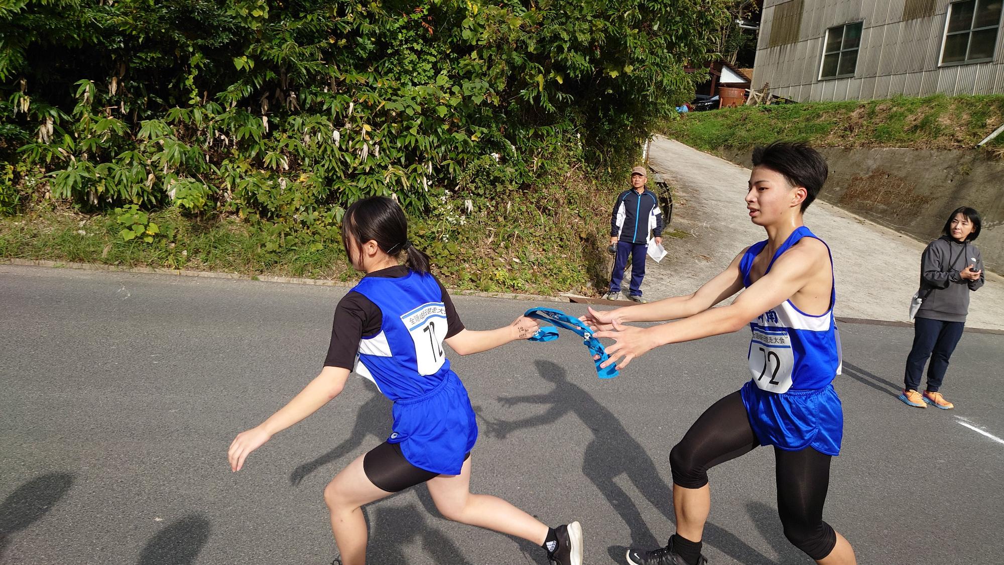 道路で男子ランナーが女子ランナーにバトンを渡している駅伝競走の写真