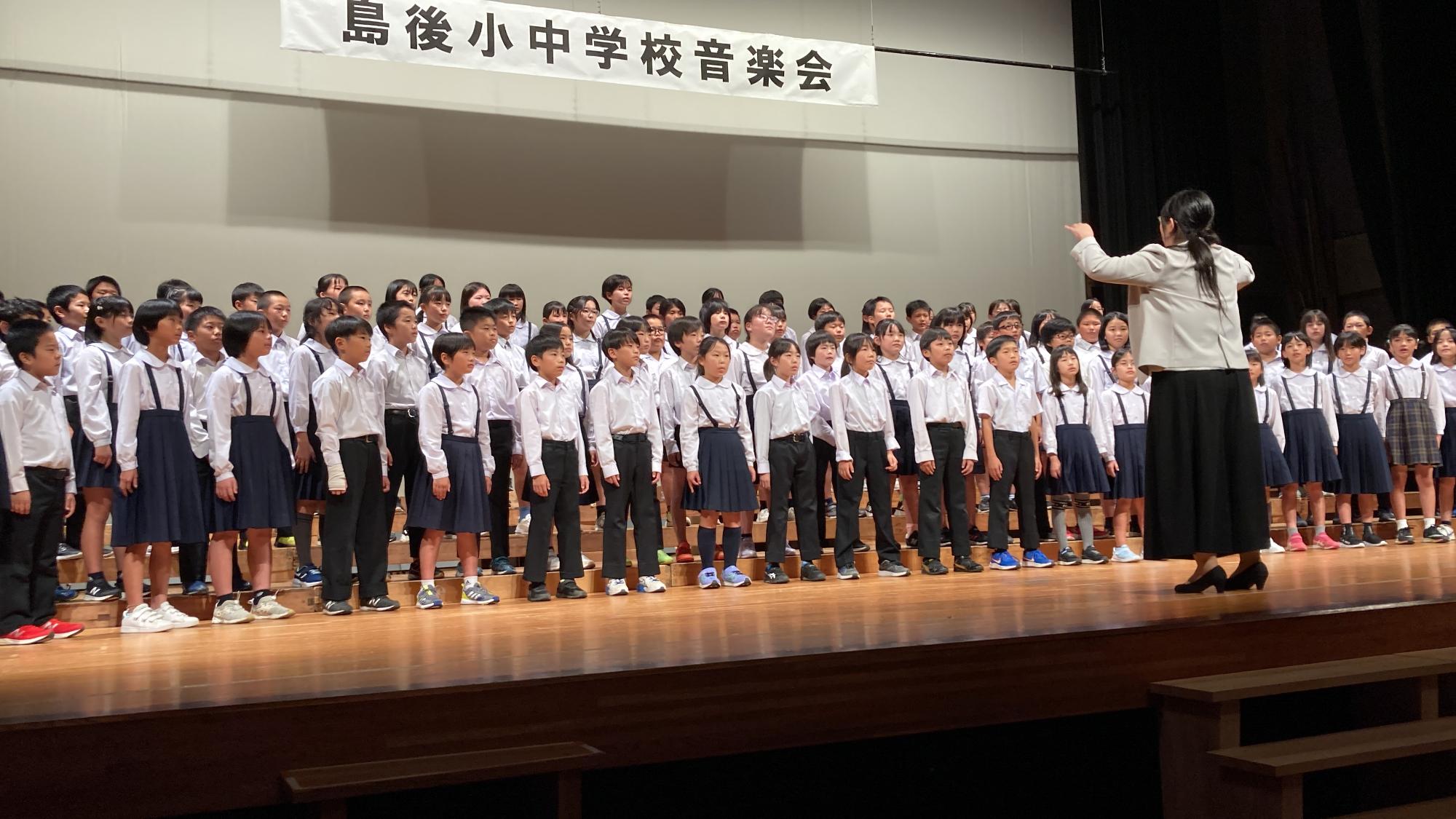 舞台上で児童たちが指揮者に合わせて合唱している写真