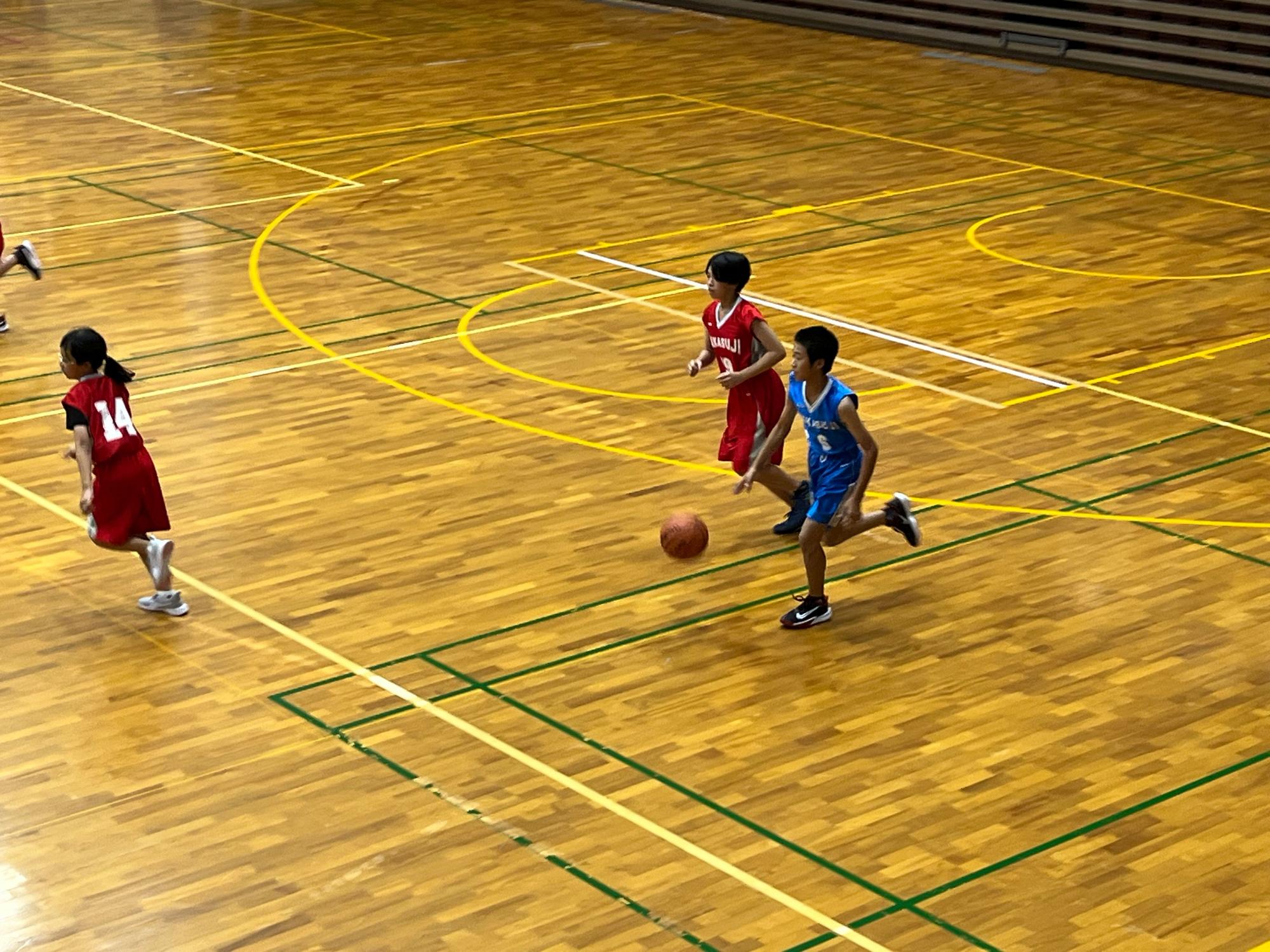 体育館でバスケットボールの試合をする赤色や青色のユニフォームを着た児童たちの写真