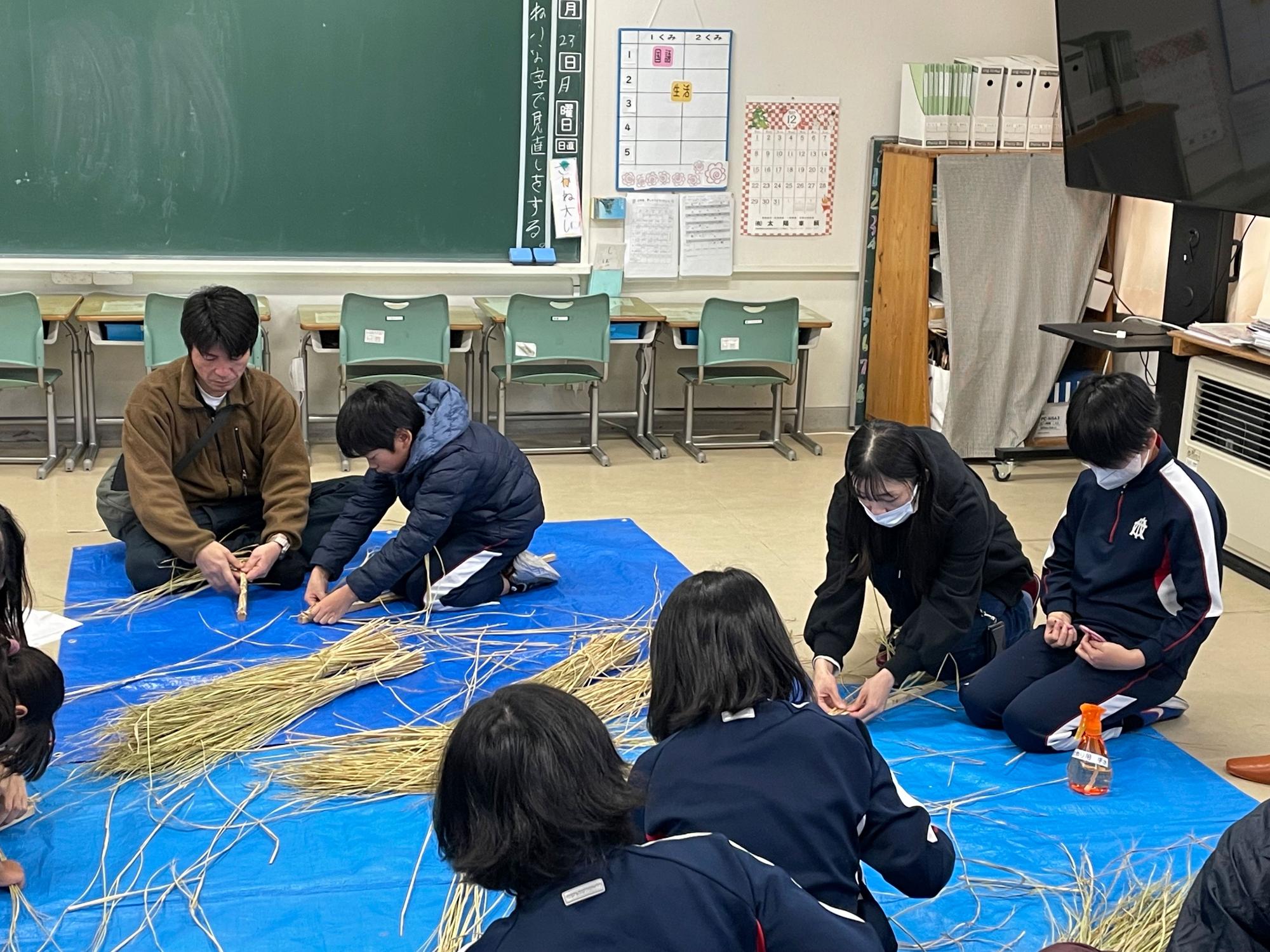 教室でブルーシートを敷きしめ縄づくりを体験する児童と保護者の写真