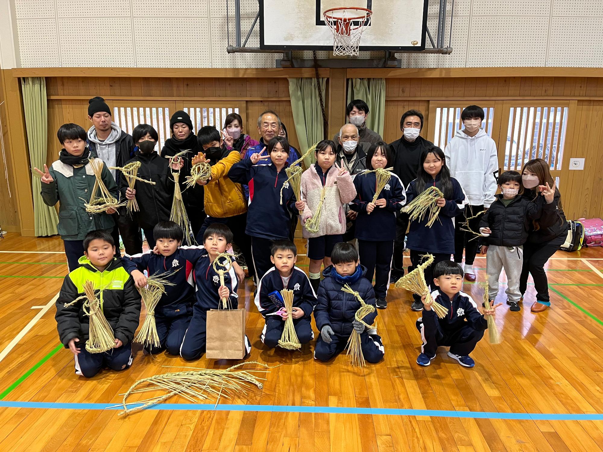 体育館で完成したしめ縄を持って集合写真を撮る児童と地域の人の集合写真