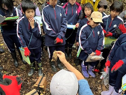 森の中で講師の話を聞くジャージ姿の児童たちの写真