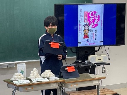 教室の黒板の前でモニターを使いながら特産品について発表する児童の写真