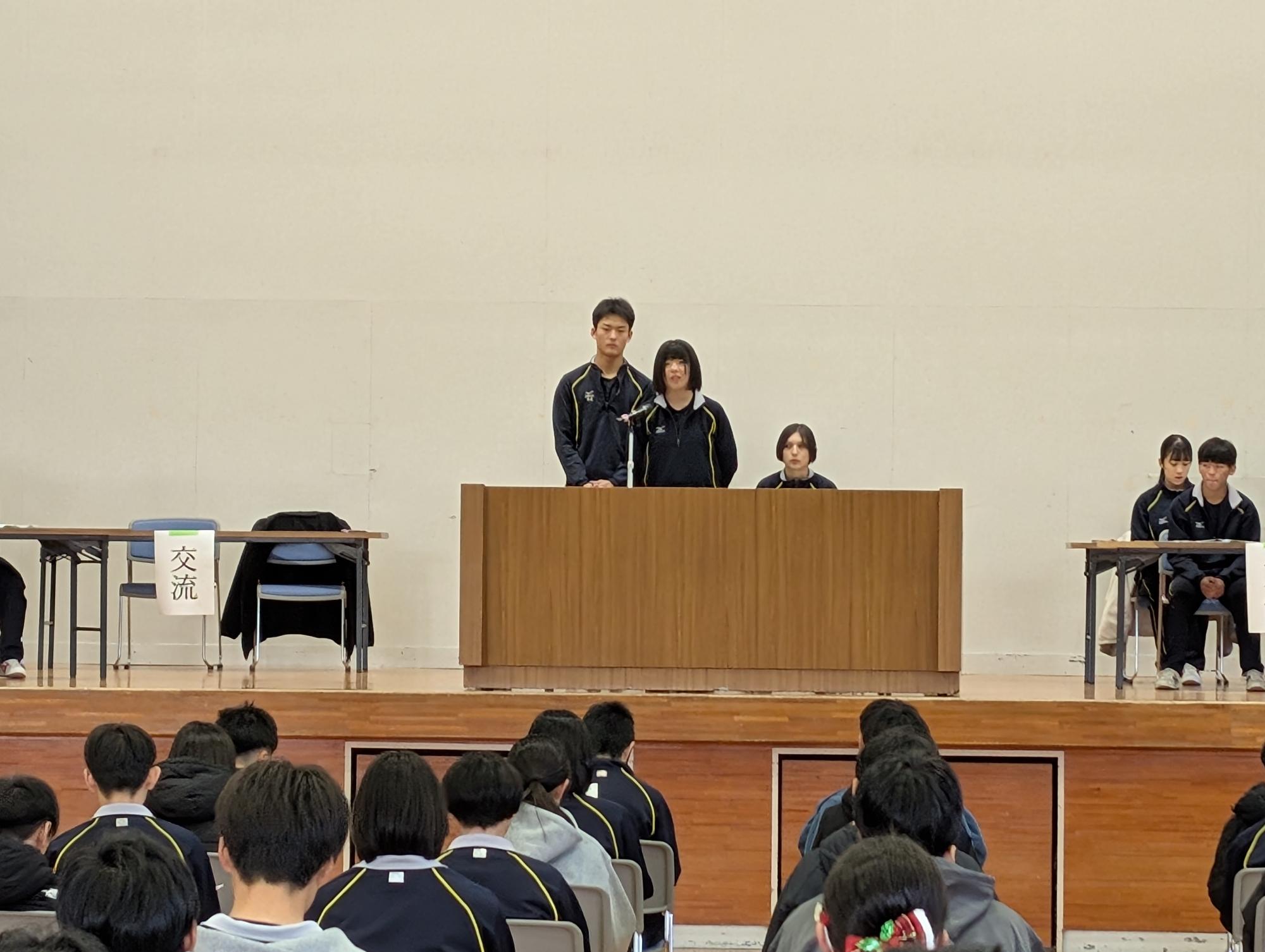 体育館の壇上で生徒会の代表が発表を行い、生徒たちが話を聞いている写真