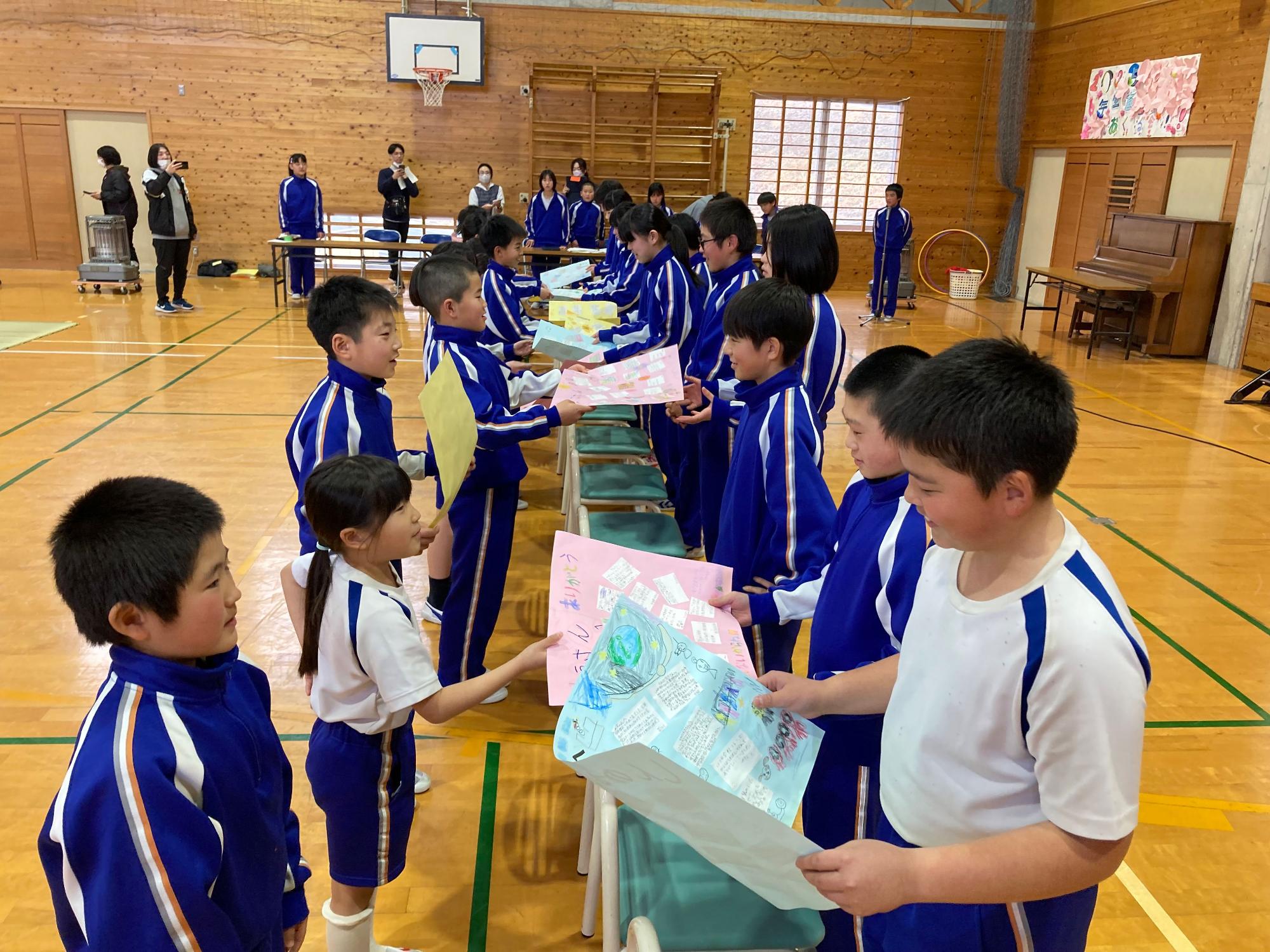 体育館で児童たちが色紙を手に持って高学年の生徒に感謝を伝えている写真