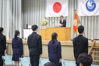 式典会場で校旗と国旗の前に生徒が整列し、壇上の先生が話をしている写真