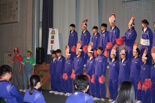 体育館の舞台で青いジャージ姿の生徒たちが演劇をしている写真