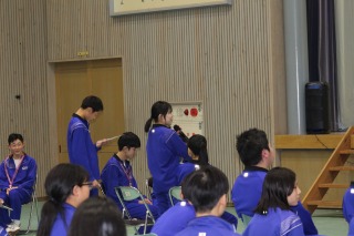 体育館で座っている生徒の前でマイクを持って発表している女子生徒の写真