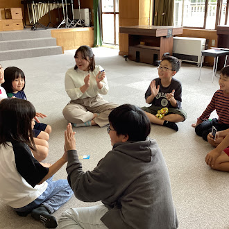 教室の床に座って先生と一緒に手遊びをしている児童たちの写真