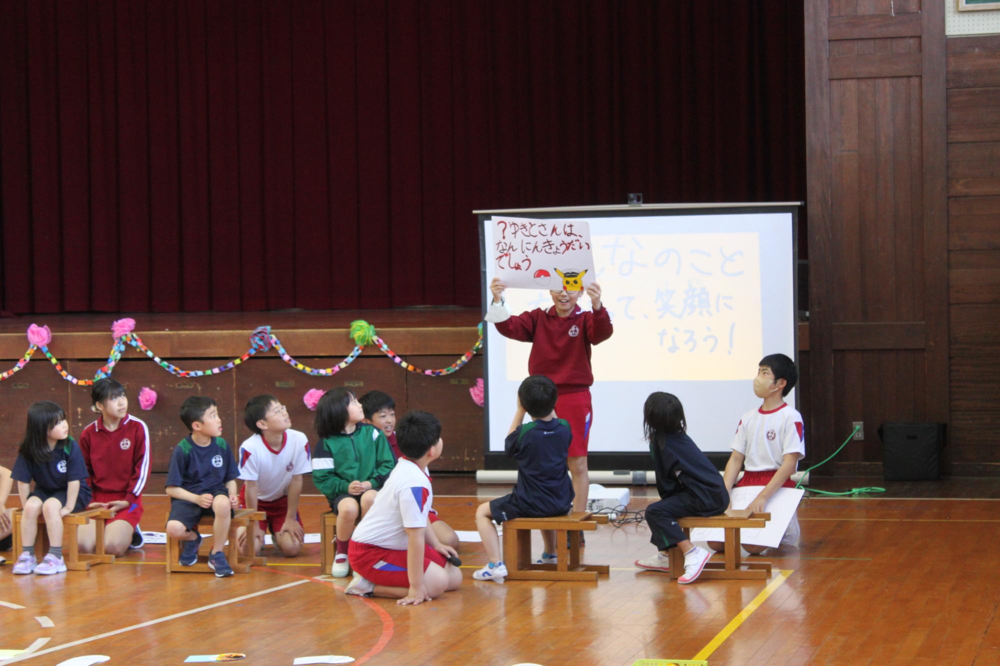 体育館で生徒がスクリーンを用いながら発表を行い、複数の生徒がそれを見ている写真