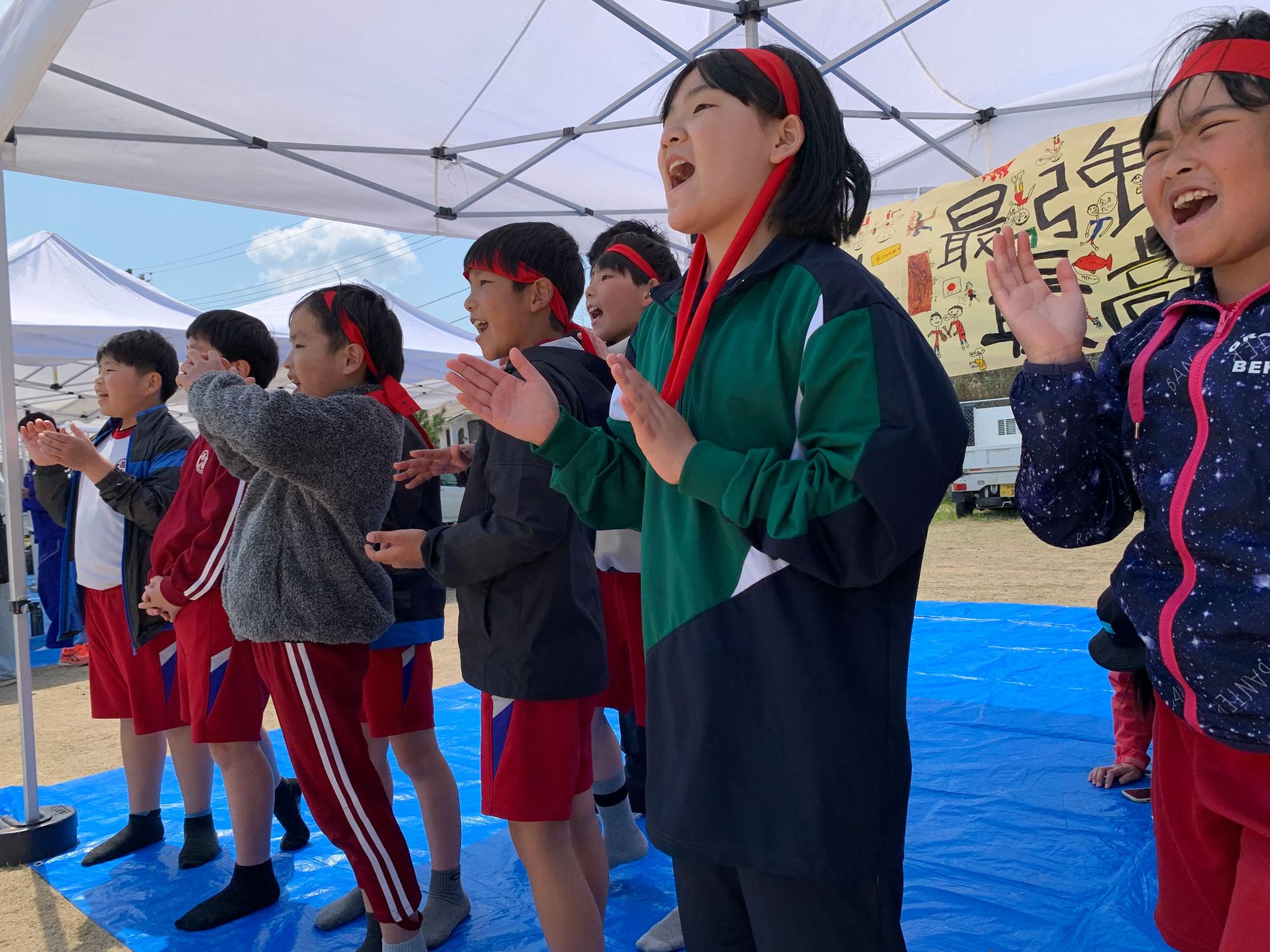 テントの下で赤いはちまきをした児童たちが応援している写真