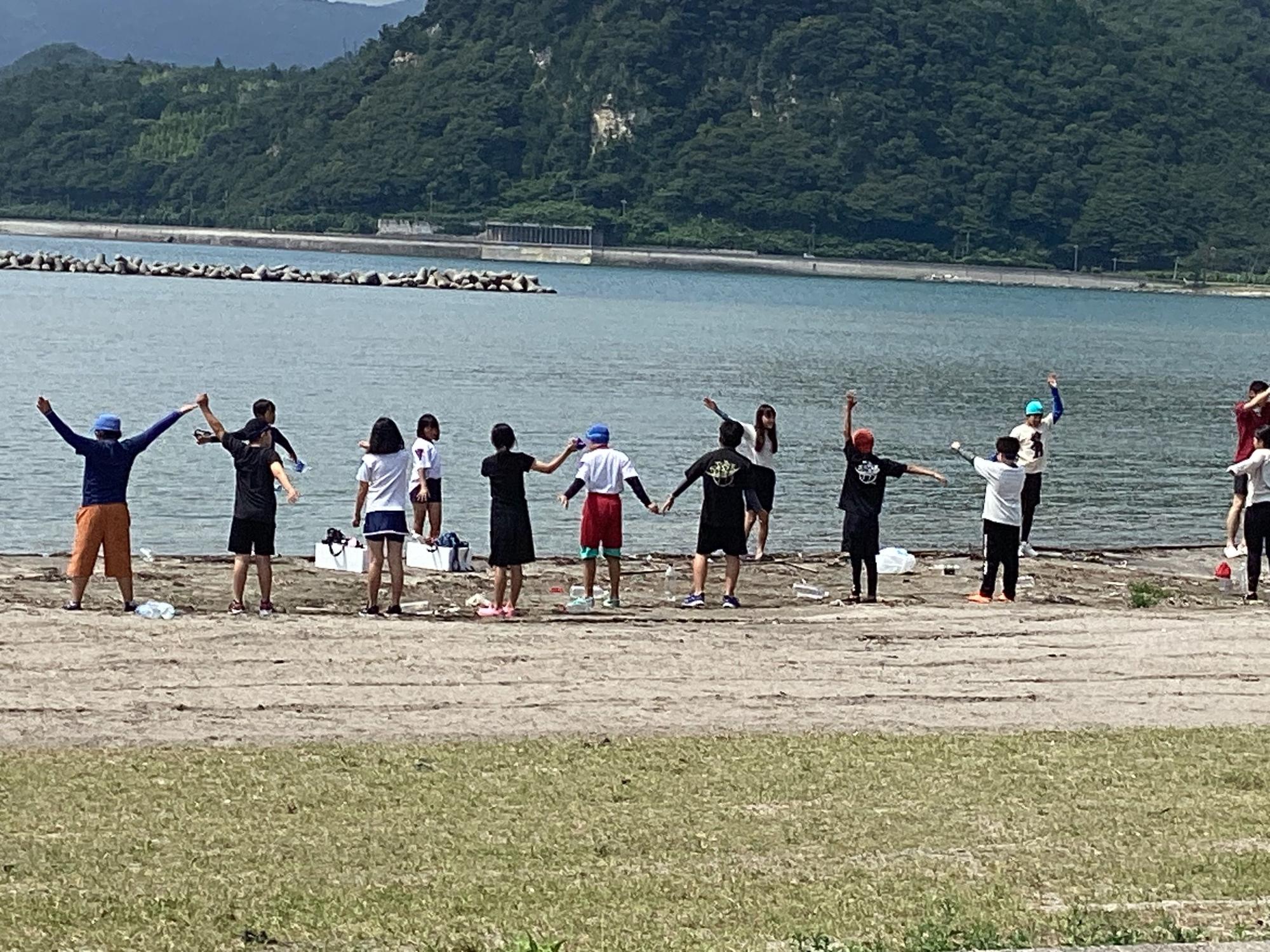 海辺の砂浜で手を広げて準備運動をするたくさんの生徒たちの写真
