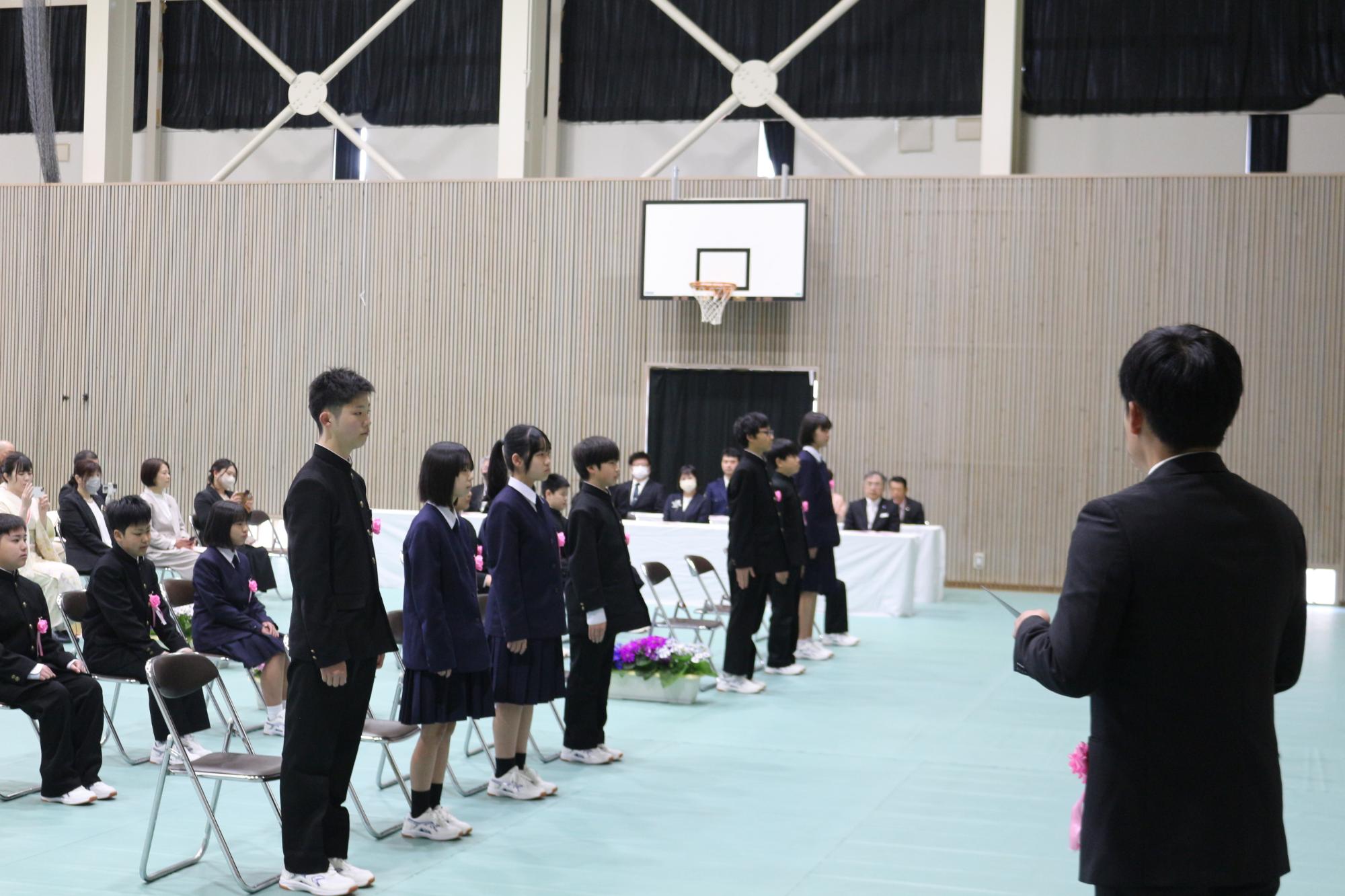 体育館で生徒たちが起立しそれを教員が見守っている写真