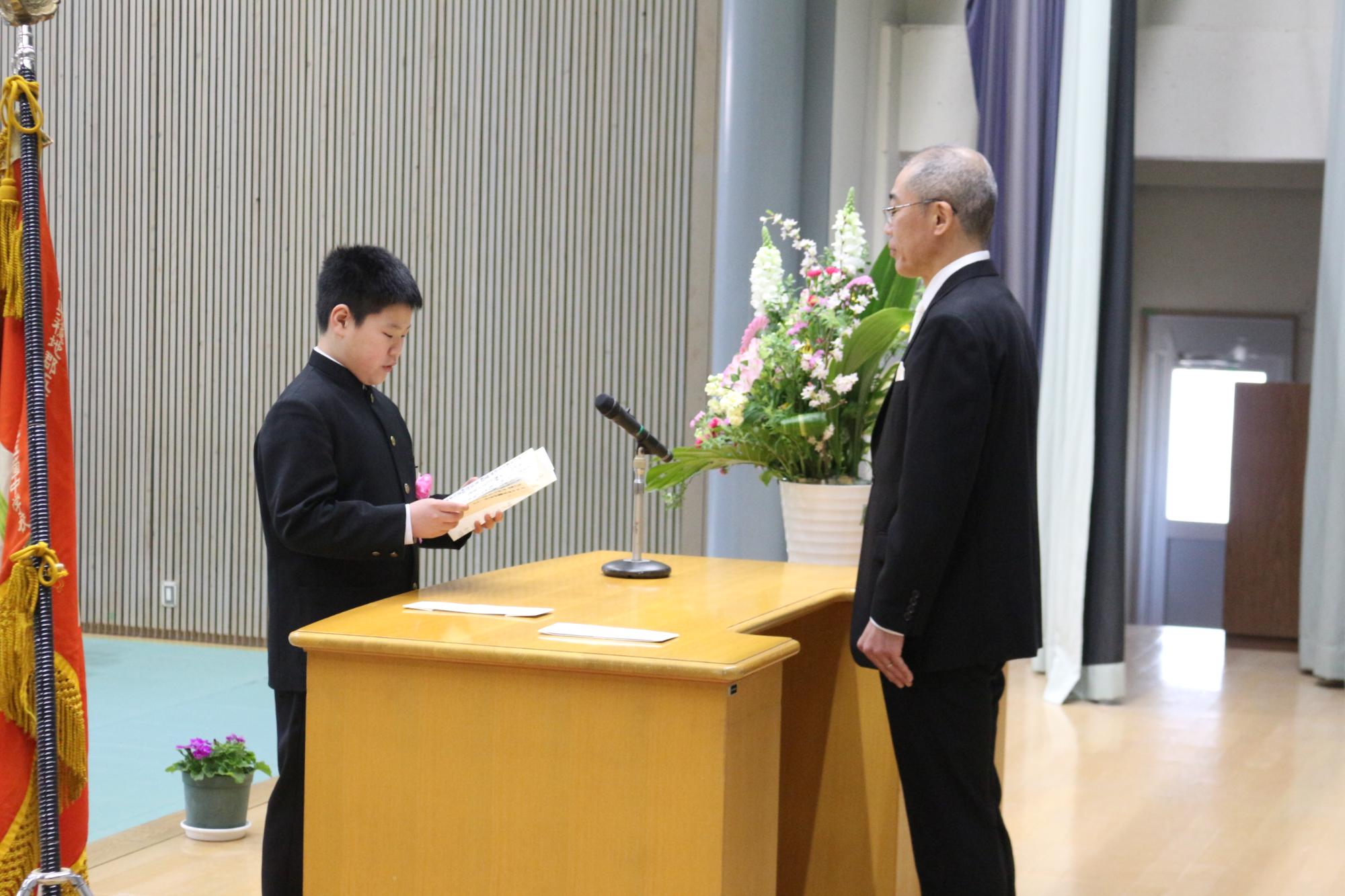 入学式で男子生徒が壇上に上がり男性教員の前で証書を読み上げている写真