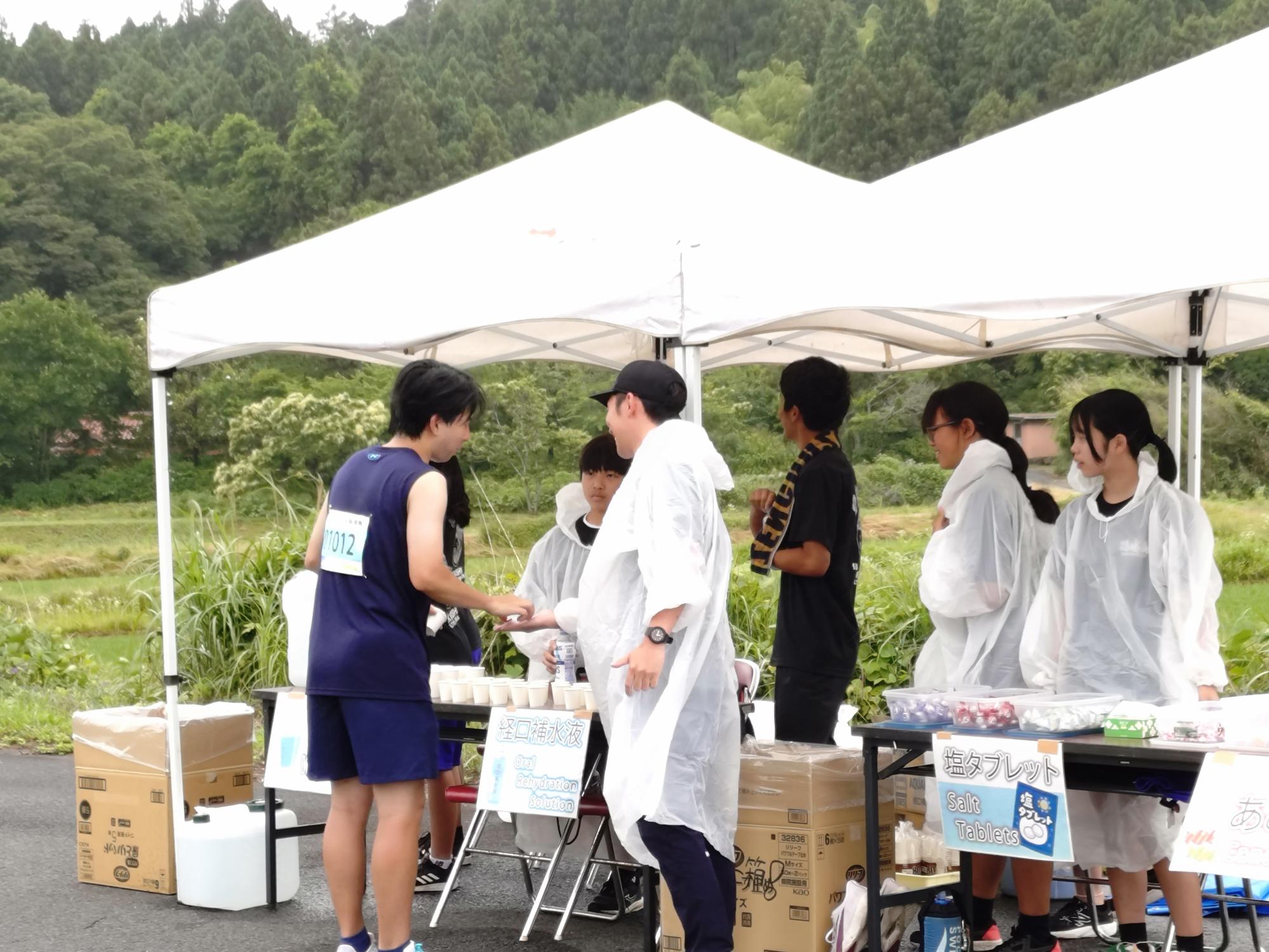 テントの下で雨具を着た生徒たちがランナーに飲み物を手渡している写真