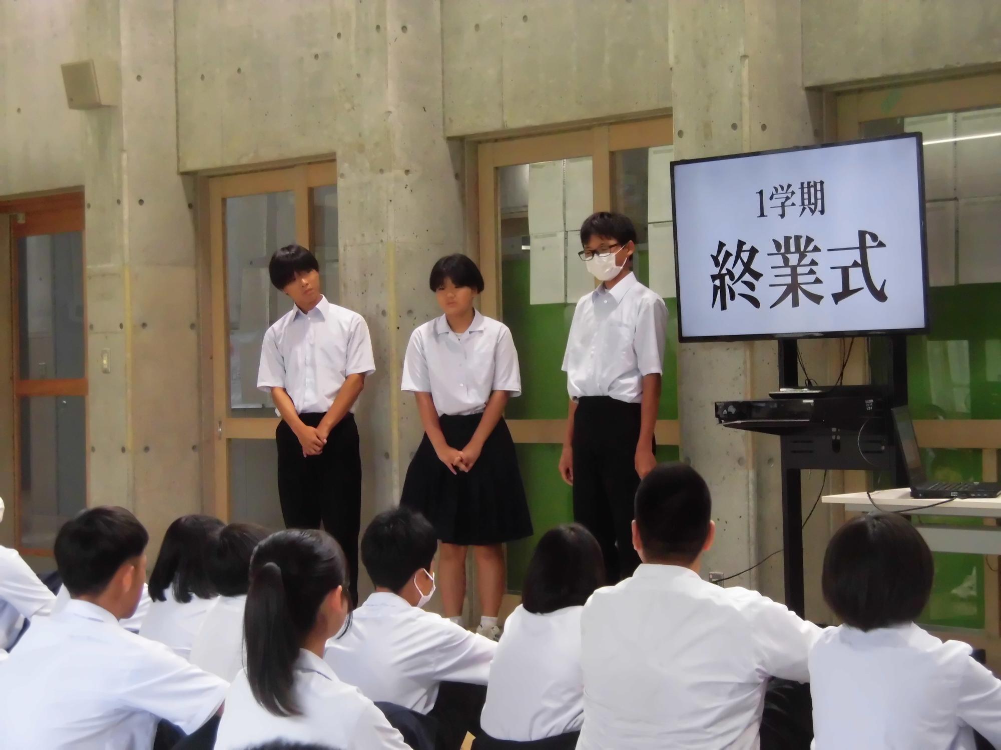 教室で1学期終業式と表示されたモニターの前で3人の生徒が立っている写真